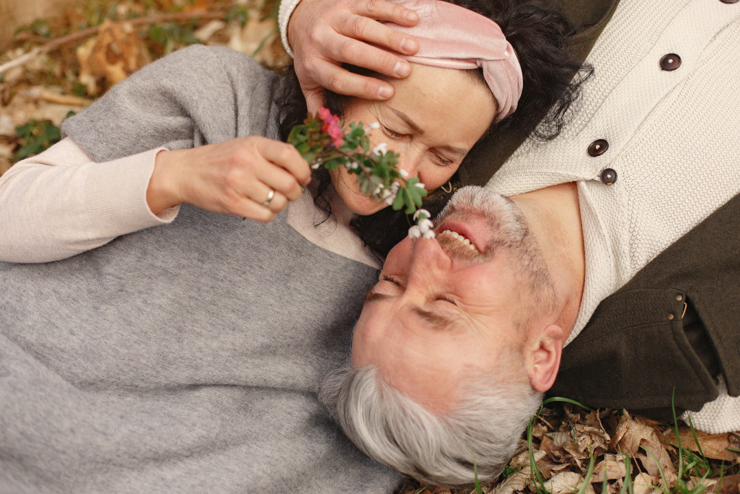 Ein älteres Paar liegt auf dem Herbstlaub auf dem Rücken. Sie spielt liebevoll mit einem Blütenstängel an seiner Nase herum.